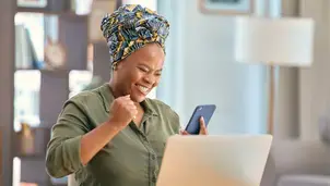 A joyful woman in a head wrap fist pumps while looking at her smartphone in front of a laptop